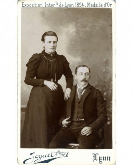 Couple: femme debout, main sur l'épaule de l'homme moustachu assis