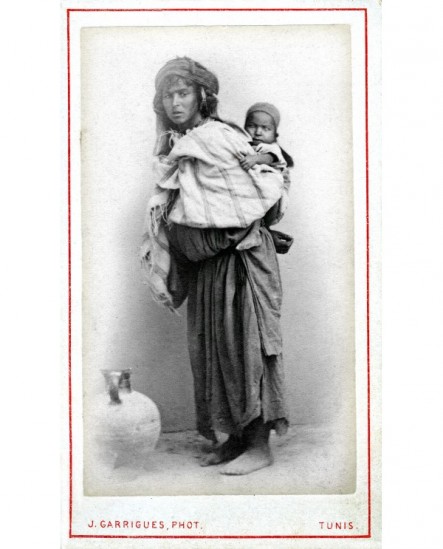 Femme tunisienne un enfant dans le dos, cruche posée à terre.
