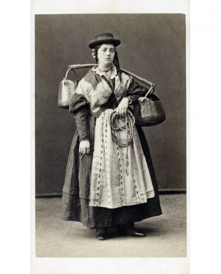 Femme en costume traditionnel, coiffée d'un chapeau masculin, debout, un jouquet avec deux chaudrons