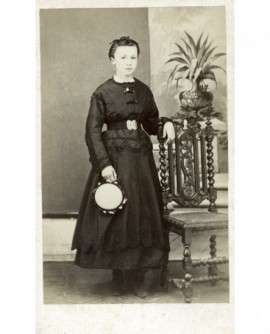 Jeune fille en robe noire, un tambourin à la main, accoudée à une chaise