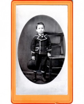 Jeune garçon en costume bordé d'un galon debout, chapeau en main