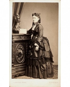 Femme en robe à tournure debout contre un meuble, tenant un livre